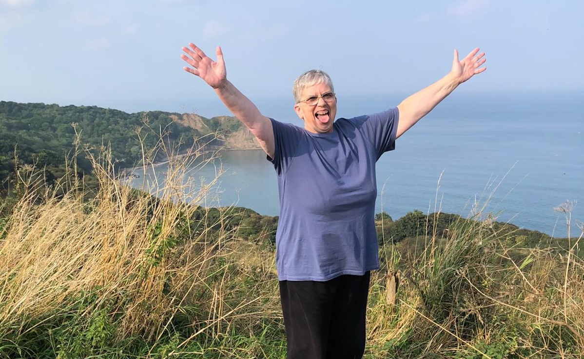 happy person with their arms in the air facing the camera standing at grassy cliffs with the sea going off into the distance behind them