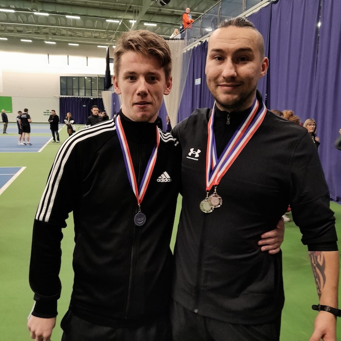 alt=“two happy proud young men in a sports centre with medals around their necks and an arm around each other facing the camera