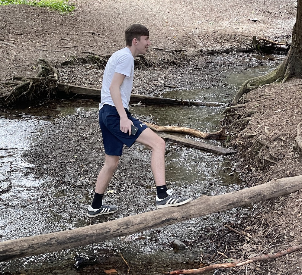 man wearing shorts and a t shirt walking on a log that is over a small river