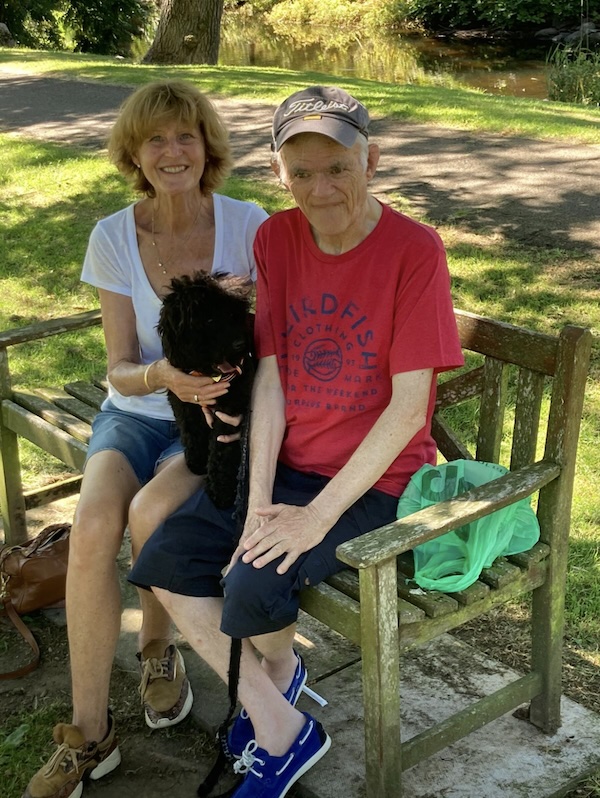 two people on a bench in a park on a warm summers day with a small black dog