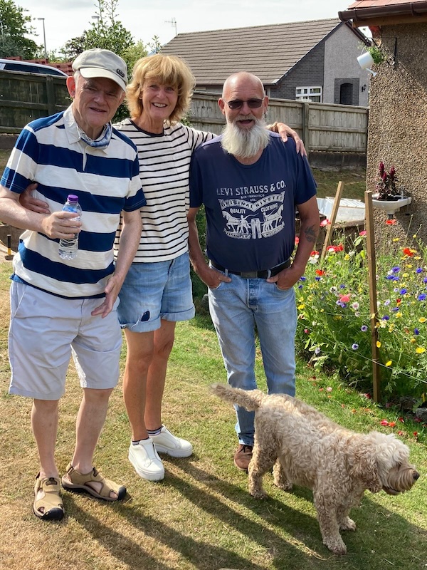 three happy people standing in a garden with a mall white dog